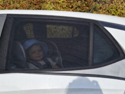 Baby in rear car seat behind Qualizzi mesh sunshade with window partially open for airflow and UV protection